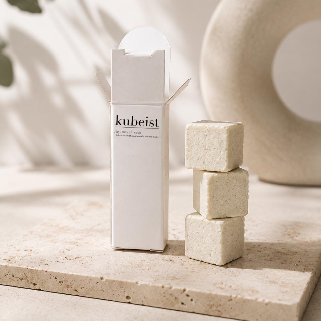 A white rectangular product box labeled 'kubeist' stands beside three stacked beige cube-shaped bath or body bars on a light stone surface, with soft neutral decor in the background.