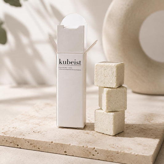A white rectangular product box labeled 'kubeist' stands beside three stacked beige cube-shaped bath or body bars on a light stone surface, with soft neutral decor in the background.